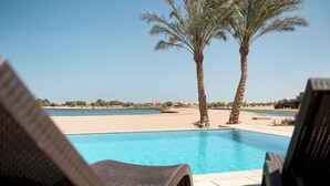 A poolside area with lounge chairs, a palm tree, and a view of the beach and distant buildings.