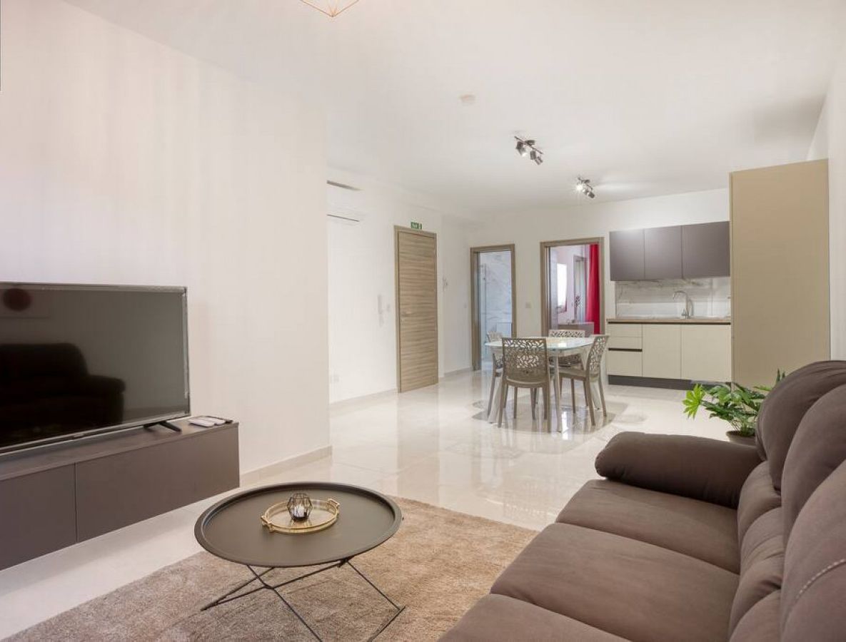 A modern living room with a flat-screen TV, a coffee table, and a sofa. There is a dining area with a table and chairs, and a kitchen in the background.