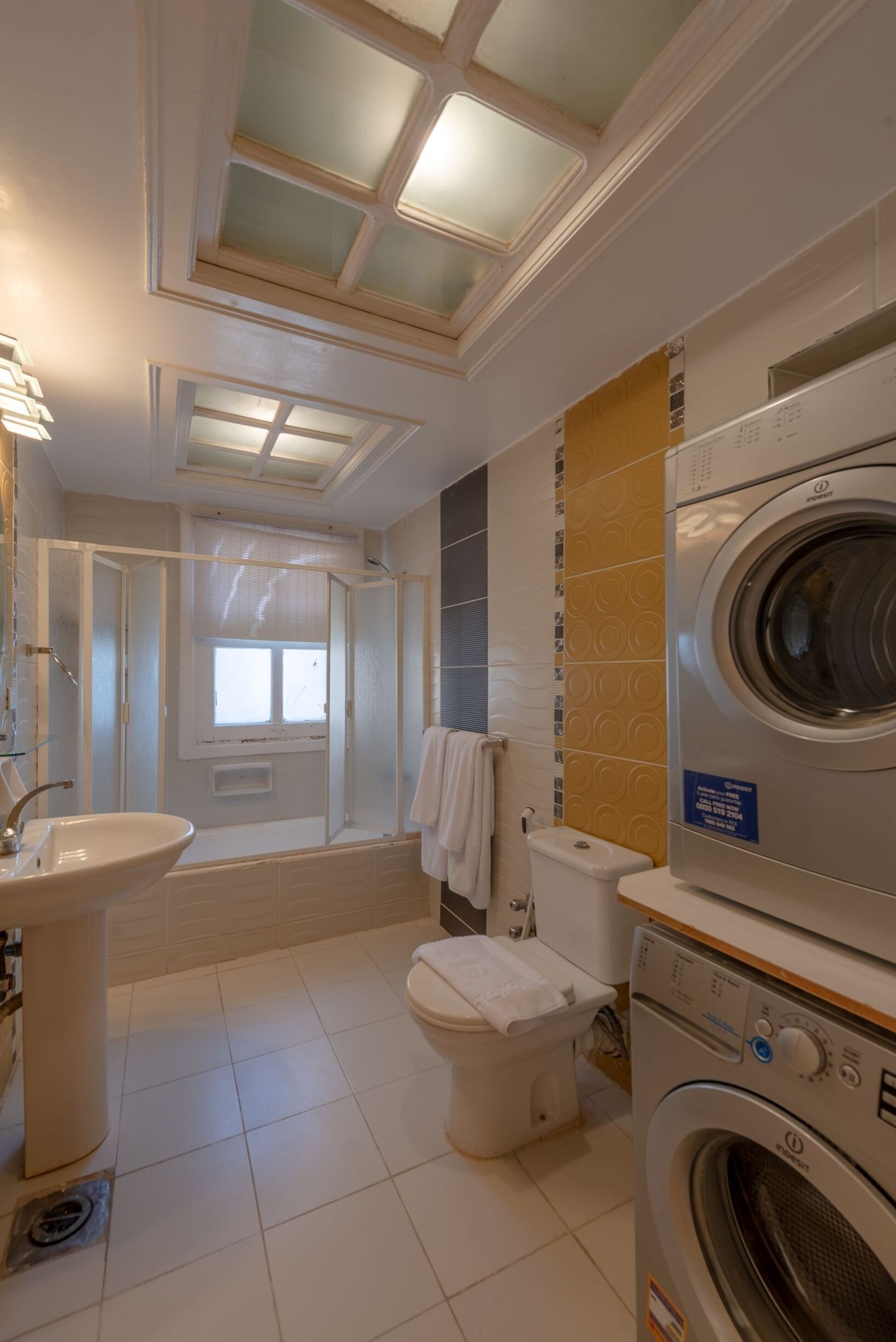Une salle de bain moderne dotée d’un puits de lumière, d’un lave-linge et d’un sèche-linge intégrés, de toilettes et d’un lavabo.