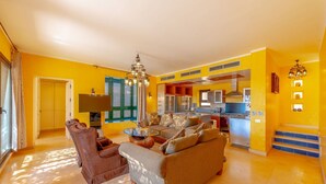 Un salon lumineux avec un canapé, des fauteuils et une table basse. Au fond, on aperçoit un coin cuisine avec une cuisinière et des placards.