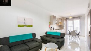 Ein modernes Wohnzimmer mit zwei grauen Sofas und einem kleinen weißen Tisch, eine Küchenzeile mit Schränken und einem Esstisch, sowie ein Gemälde von einem Leuchtturm.