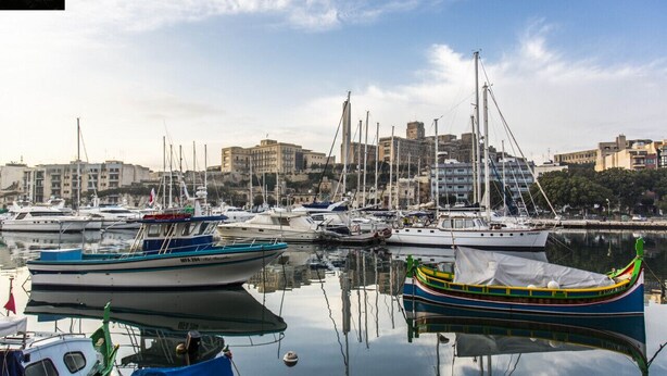 Una marina con diverse imbarcazioni ormeggiate, uno skyline cittadino sullo sfondo e cielo sereno.