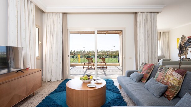 Ein Wohnzimmer mit einem blauen Shaggy-Teppich, einem Couchtisch mit Obst, einem grauen Sofa mit bunten Kissen und einem Fernseher auf einer Holzkommode.