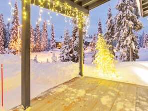 Un paisaje nevado con una terraza de madera, muñecos de nieve y un árbol de Navidad iluminado con luces.
