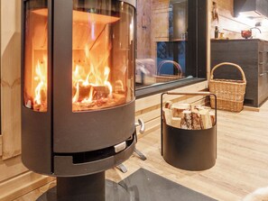 Un poêle à bois avec une façade en verre, un panier de bois de chauffage et un coin cuisine doté de meubles en bois et d’une fenêtre.