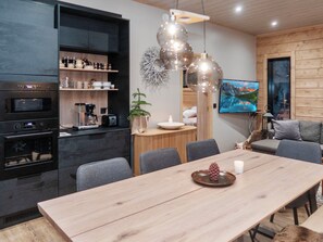Une cuisine moderne dotée d’appareils électroménagers intégrés, un coin repas avec une table en bois et un espace salon confortable avec un téléviseur diffusant un paysage de montagne.