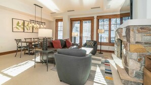 A spacious living room with a stone fireplace, large windows, and a modern chandelier. - Big Horn Lodge 410 - Platinum Slopeside Residence (Truckee)