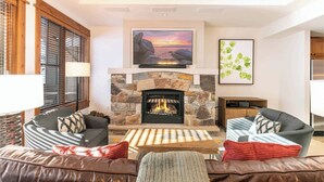 A living room with a stone fireplace, two sofas, a coffee table, and a large painting of a sunset. - Big Horn Lodge 410 - Platinum Slopeside Residence (Truckee)