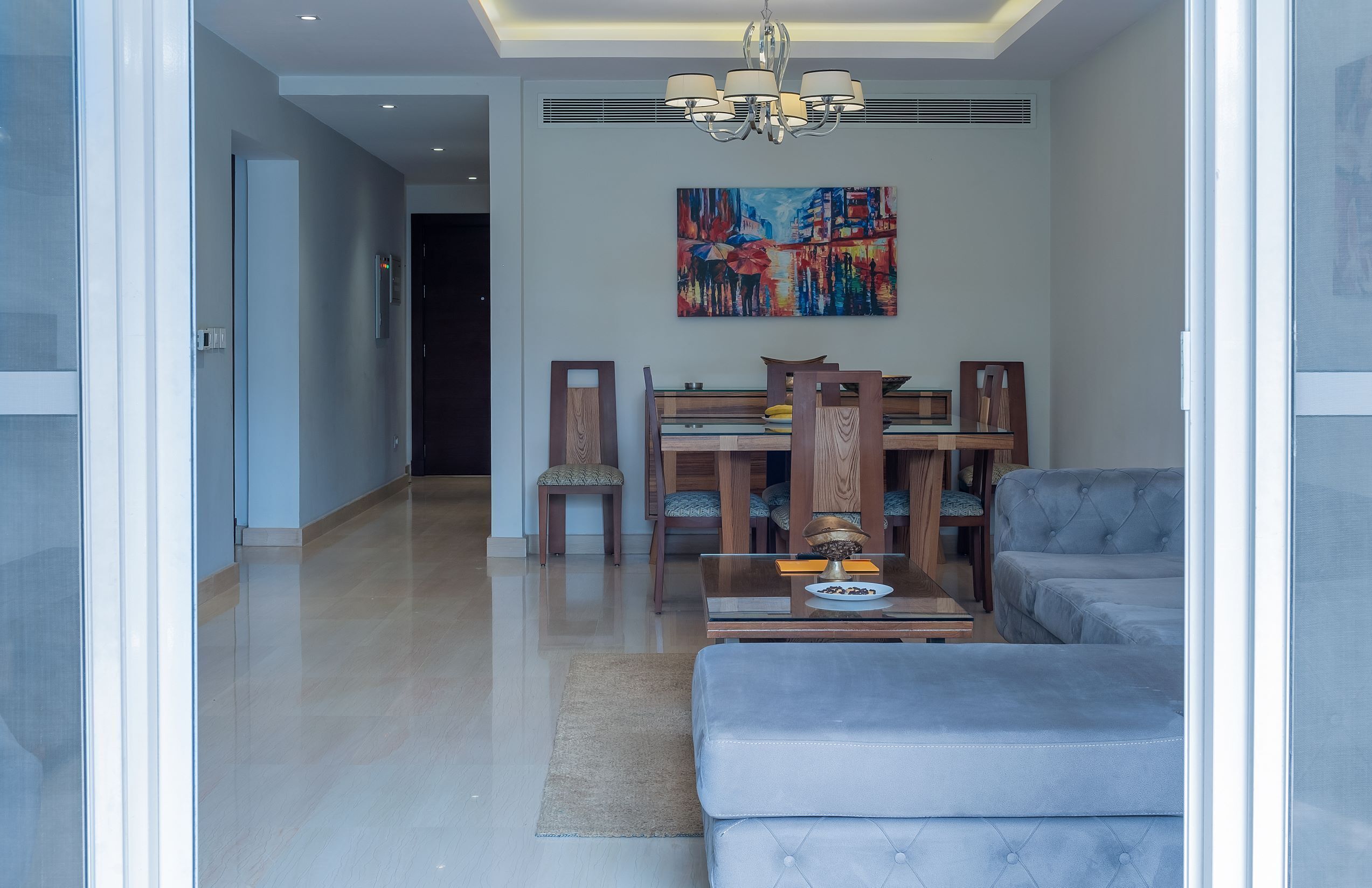 A modern living room with a sofa, a dining table with chairs, a chandelier, and a painting on the wall.