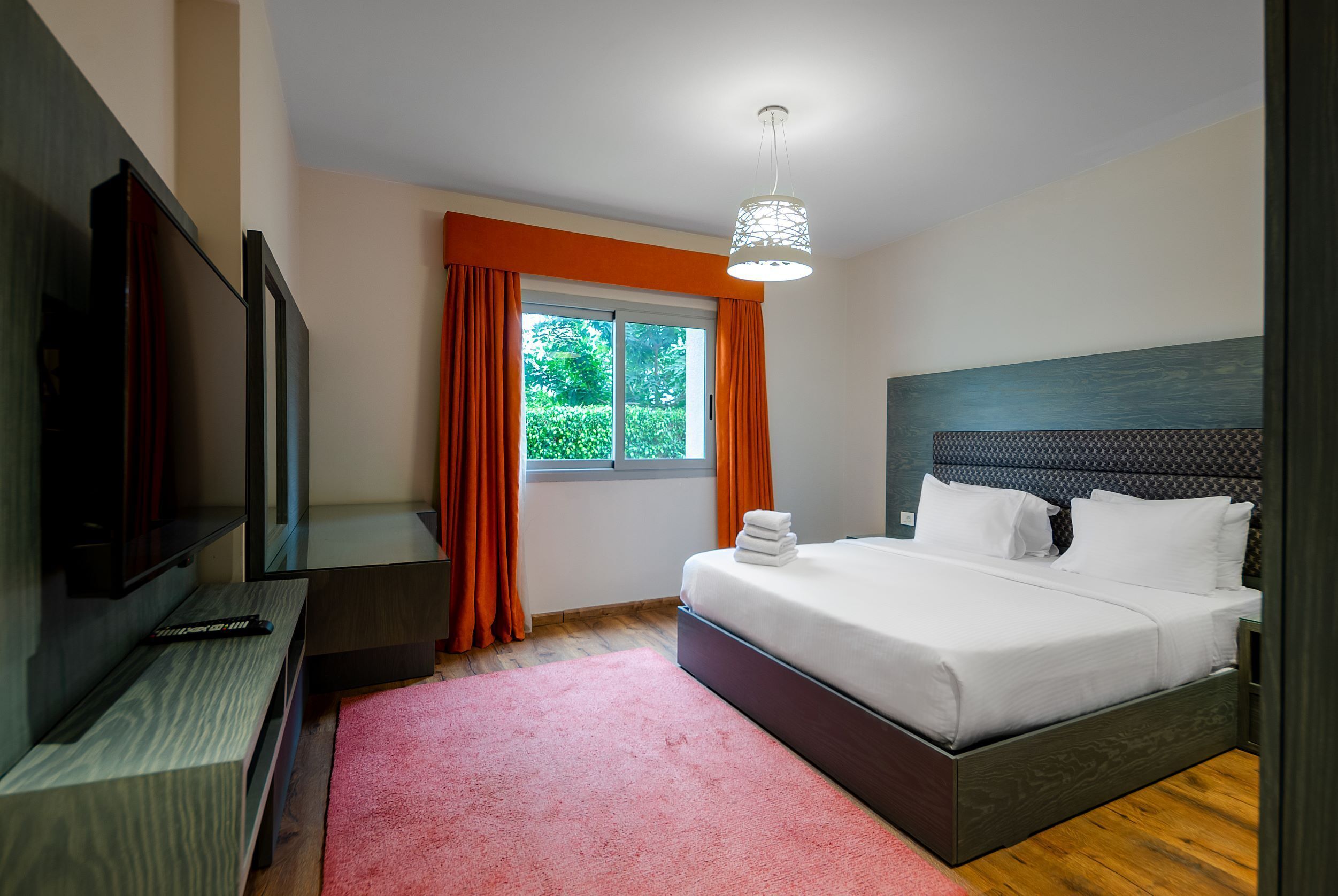 A modern hotel room with a large bed, a flat-screen TV, and a window with orange curtains.