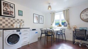 A compact kitchen with a washing machine, dining table with chairs, a clock, and a window with curtains. - Beautiful Apartment • Close to Town Centre • Free Parking • Fast-Wifi • Sky TV • By Yoko Property (Northampton)