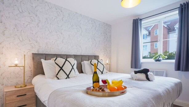 A hotel room with a bed, a bedside table with a lamp, a window with curtains, and a tray with food and a bottle. - Beautiful Apartment • Close to Town Centre • Free Parking • Fast-Wifi • Sky TV • By Yoko Property (Northampton)