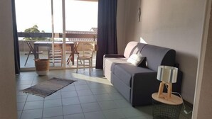 A living room with a sofa, a small table with a lamp, a basket, and a view of an outdoor patio with a table and chairs. - Studio Cabine by Interhome (Saint-Florent)