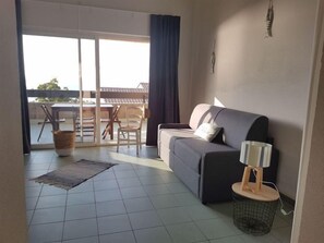 A living room with a sofa, a small table with a lamp, a basket, and a view of an outdoor patio with a table and chairs.