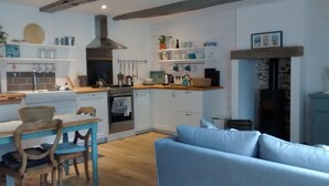 Een moderne keuken met witte kasten, een fornuis en een hangende rek voor pannen. Er staat een blauwe bank en een houten eettafel met stoelen.