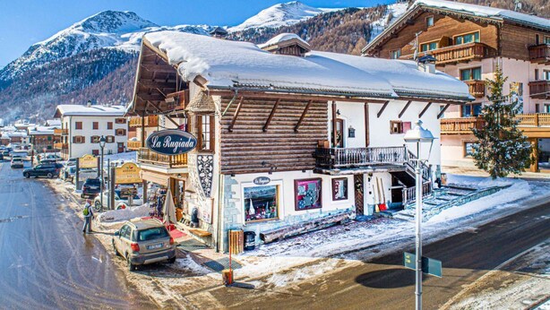 Un resort de montaña con un edificio de madera identificado como "La Rugiada", que cuenta con un balcón, una señal y vistas de montañas nevadas.