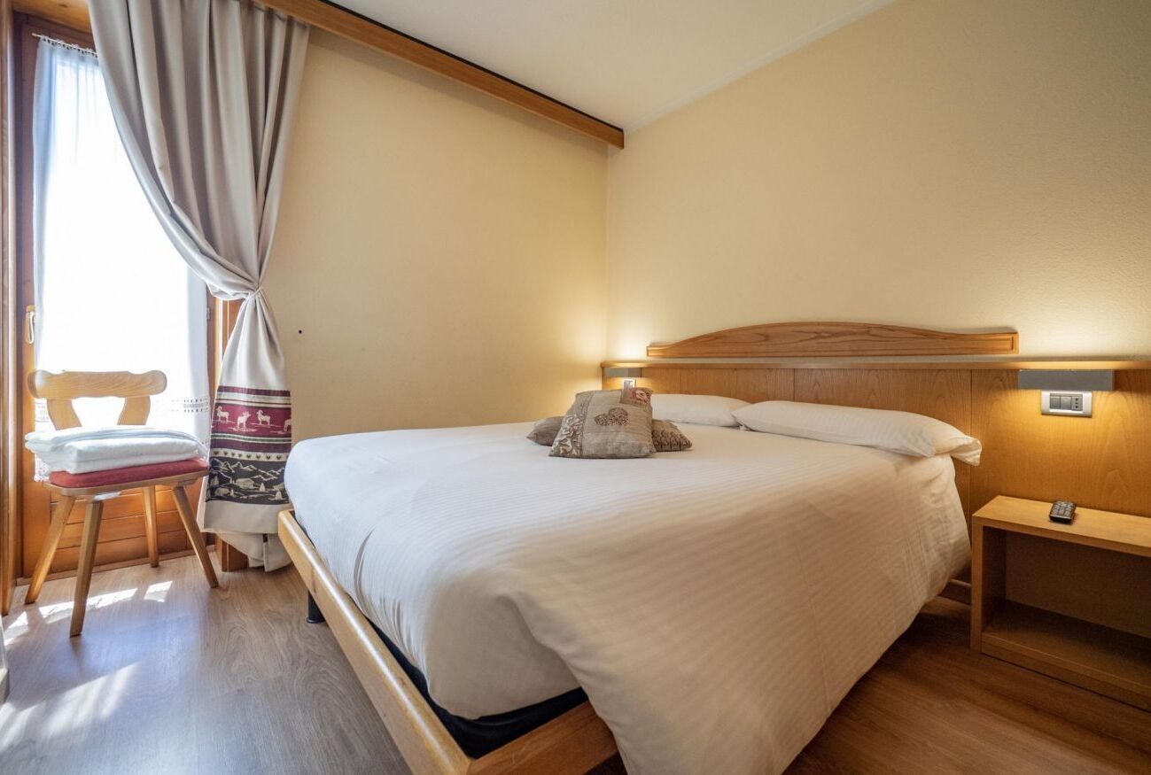 A neatly made bed with a wooden headboard, a bedside table with a remote control, and a chair with a patterned cushion.