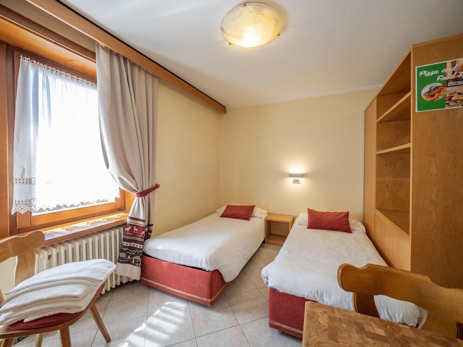 A hotel room with two single beds, a wooden chair, a radiator, a window with curtains, and a wall-mounted shelf with a menu sign.