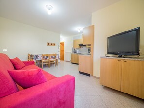 Ein kompakter Wohnbereich mit einem roten Sofa, einem Esstisch mit Stühlen, einer Küchenzeile mit Holzschränken und einem Flachbildfernseher auf einem Lowboard.