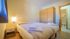 A bedroom with two beds, wooden wardrobe, bedside tables, and a bathroom visible through an open door.