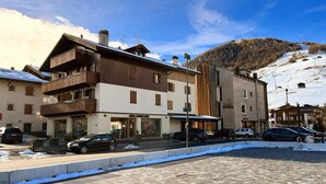 Ein mehrstöckiges Gebäude mit Holzbalkon, im Hintergrund eine Skipiste und im Vordergrund eine gepflasterte Fläche.