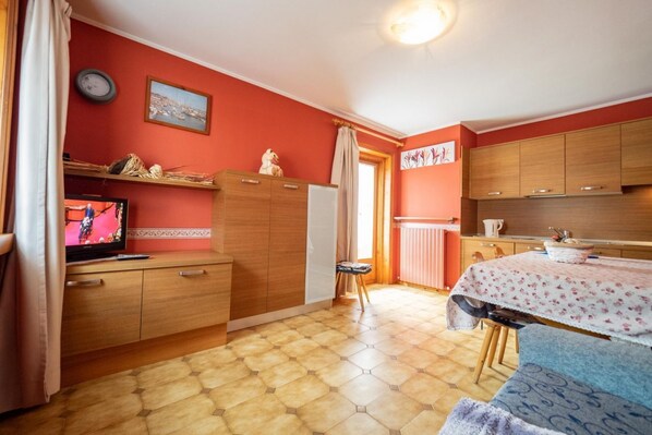 Ein kleines Zimmer mit einem Holzschrank, einem kleinen Tisch mit einer blumigen Tischdecke, einem Fernseher auf einem Holzgestell und einem Fenster mit Vorhängen.