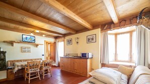 Una habitación acogedora con techo de madera, un sofá, una mesa de comedor con sillas, una zona de cocina con microondas y una ventana con cortinas.