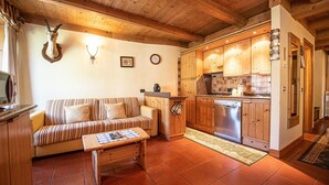Una sala de estar acogedora con un sofá a rayas, una mesa de centro de madera y una cabeza de animal montada. Junto a ella, una cocina con armarios de madera, un lavavajillas de acero inoxidable y un salpicadero de azulejos.