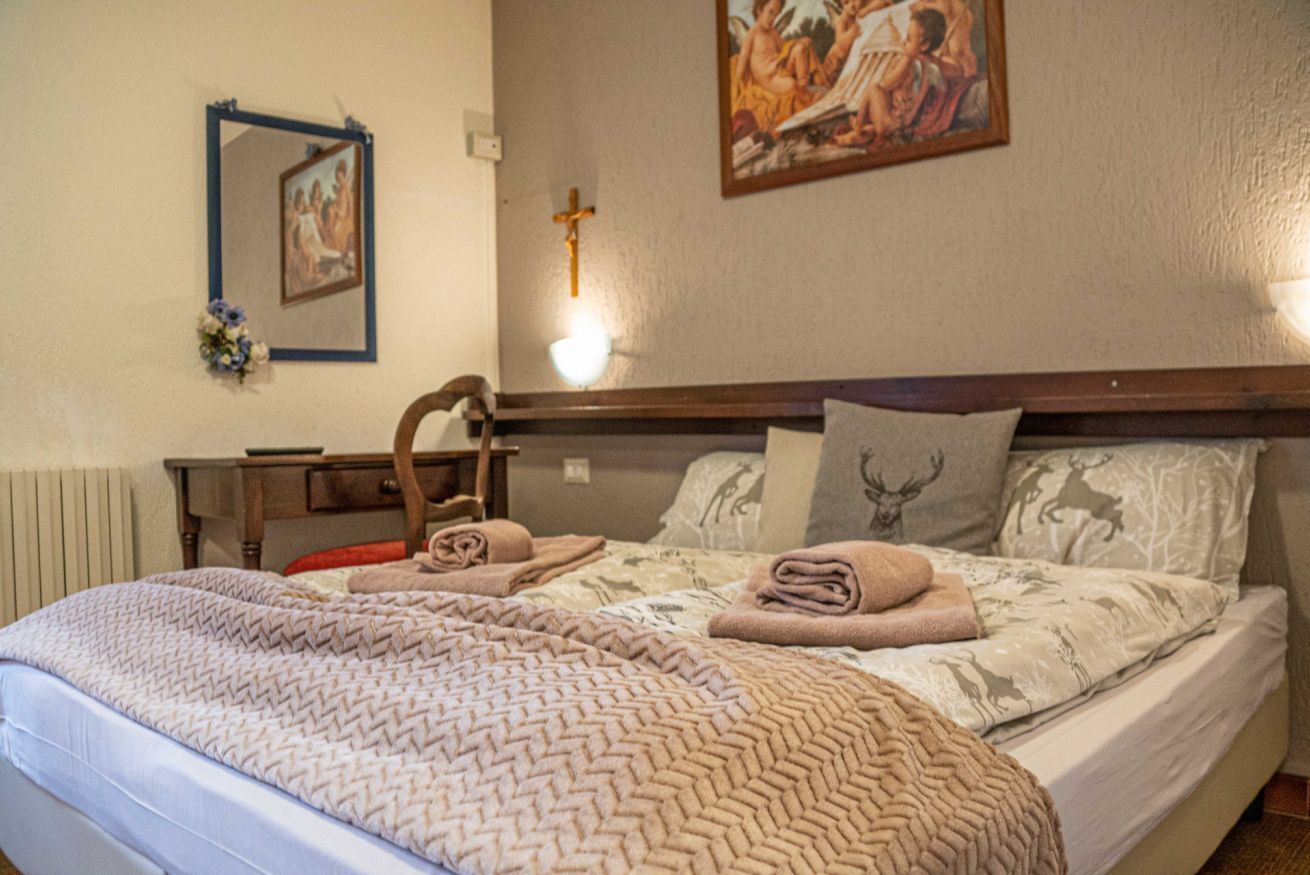 A bedroom with a bed, a nightstand, a mirror, a wall-mounted light, and a religious painting.