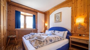 Une pièce aux murs lambrissés, comprenant un lit, une fenêtre avec des rideaux, un luminaire fixé au mur et un petit tabouret.