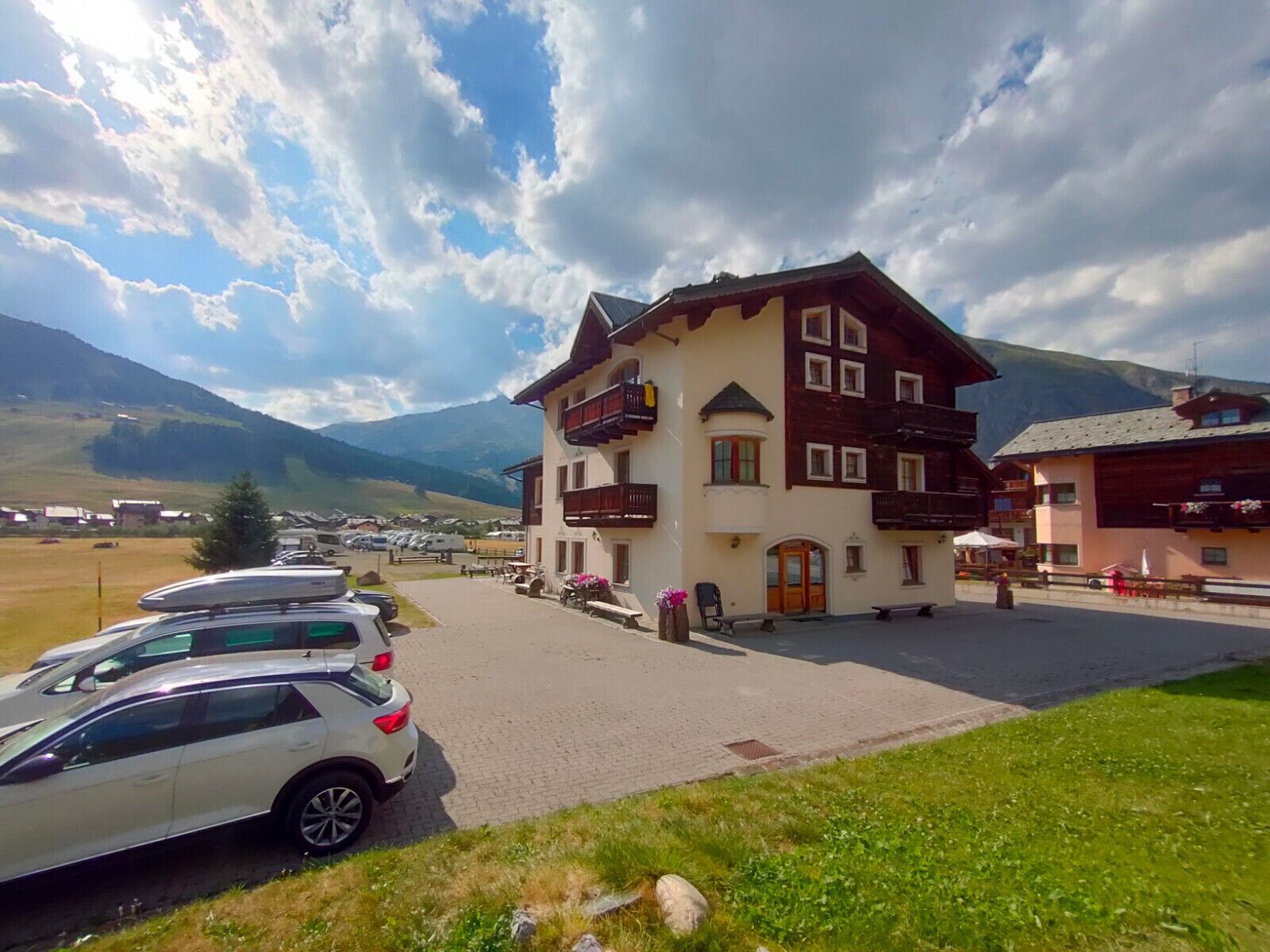 Un immeuble de deux étages doté d’un balcon, un espace de stationnement avec des voitures et un paysage montagneux en arrière-plan.