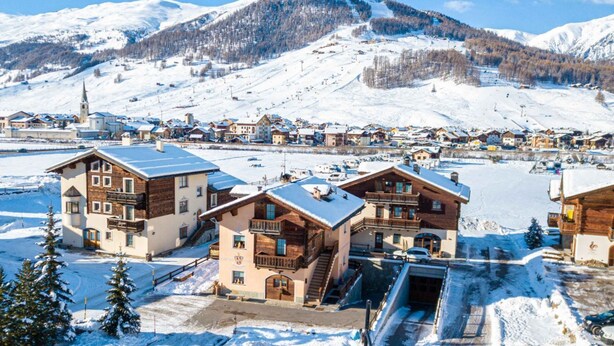 Un paese montano innevato con edifici in legno tradizionali, una seggiovia sullo sfondo e una vista chiara delle montagne circostanti.