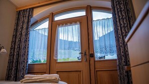 Une pièce dotée d’une porte en bois aux lignes courbes, de deux fenêtres ornées de rideaux à motifs, d’un lit recouvert d’une couvre-lit à fleurs et d’une chaise bleue.