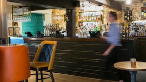 A bar area with stools, a counter with various bottles, and a blurred person in the background. - The Elephant at the Market (Newbury)