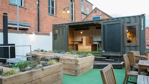 A rustic outdoor patio with wooden planters, a dining area with wooden furniture, and a brick building in the background. - The Elephant at the Market (Newbury)