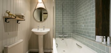 Un baño con lavamanos pedestal, un espejo redondo, una bañera y un estante con artículos de aseo.