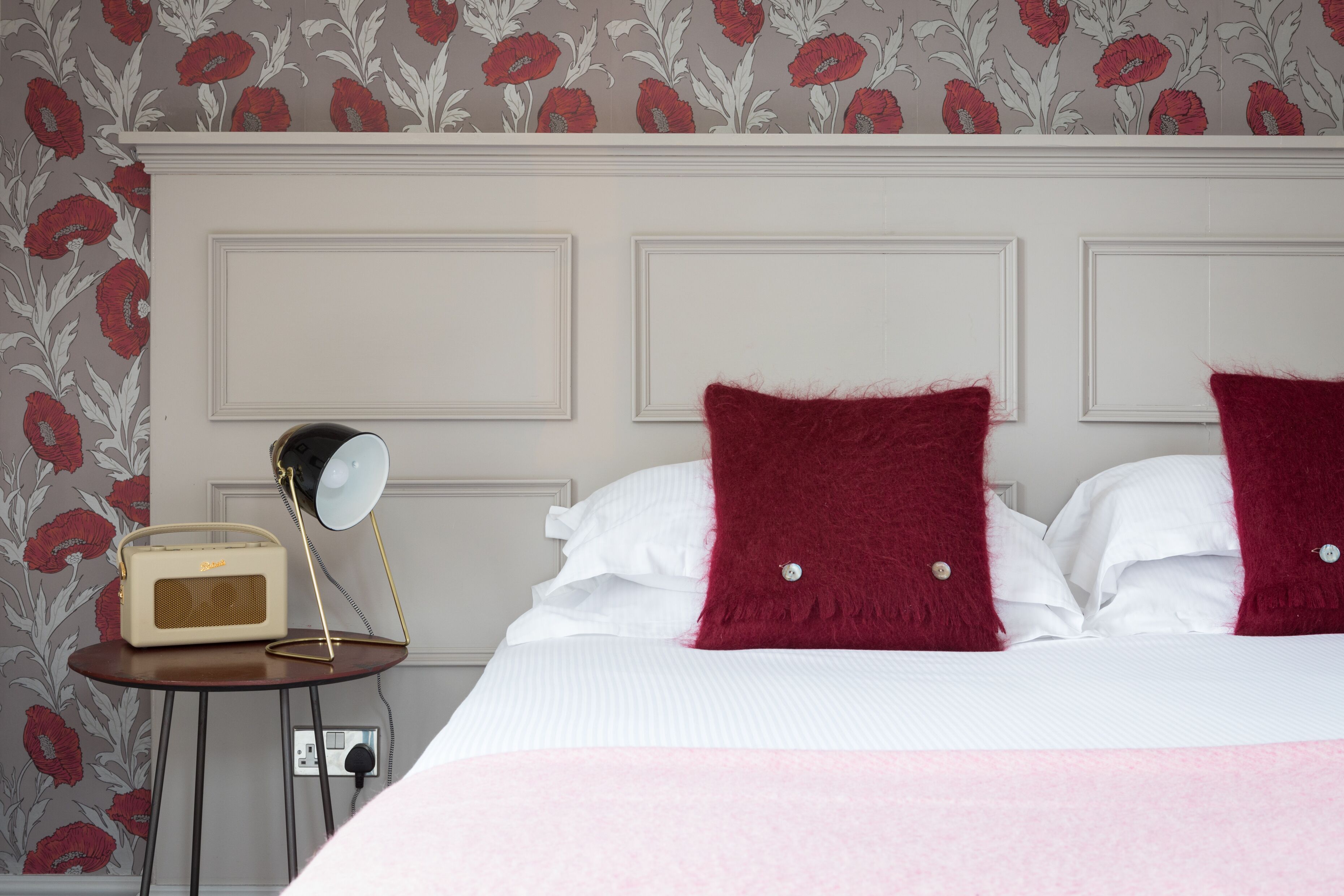 A neatly made bed with white and red pillows, a small bedside table with a lamp, and a radio.