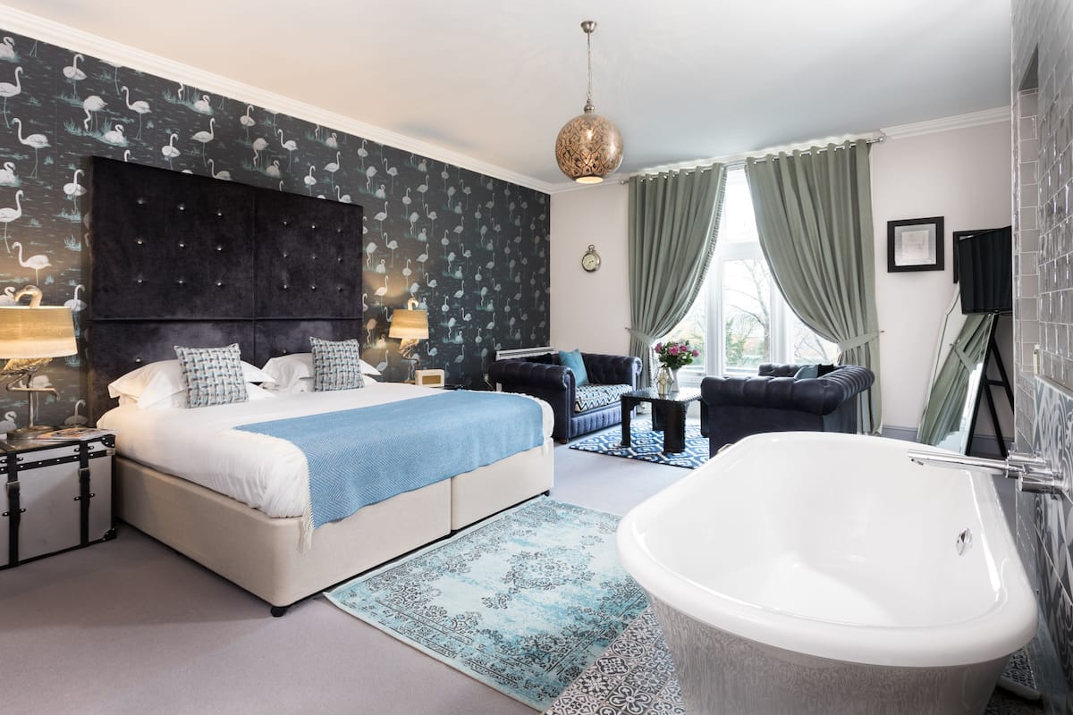 A bedroom with a large bed, a sitting area with a sofa and armchairs, a small table, a large bathtub, and a window with curtains.
