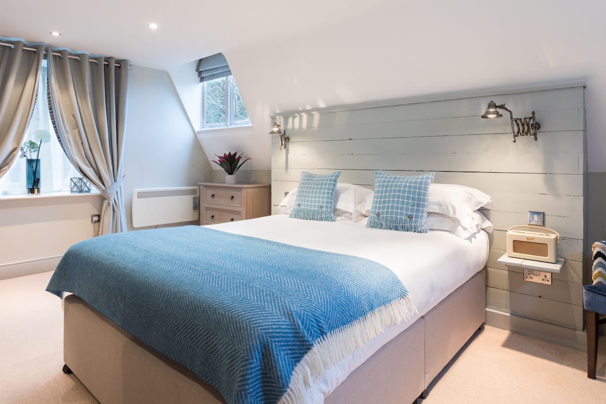 A bedroom with a large bed, a bedside table with a lamp, a small shelf, and a window with curtains.