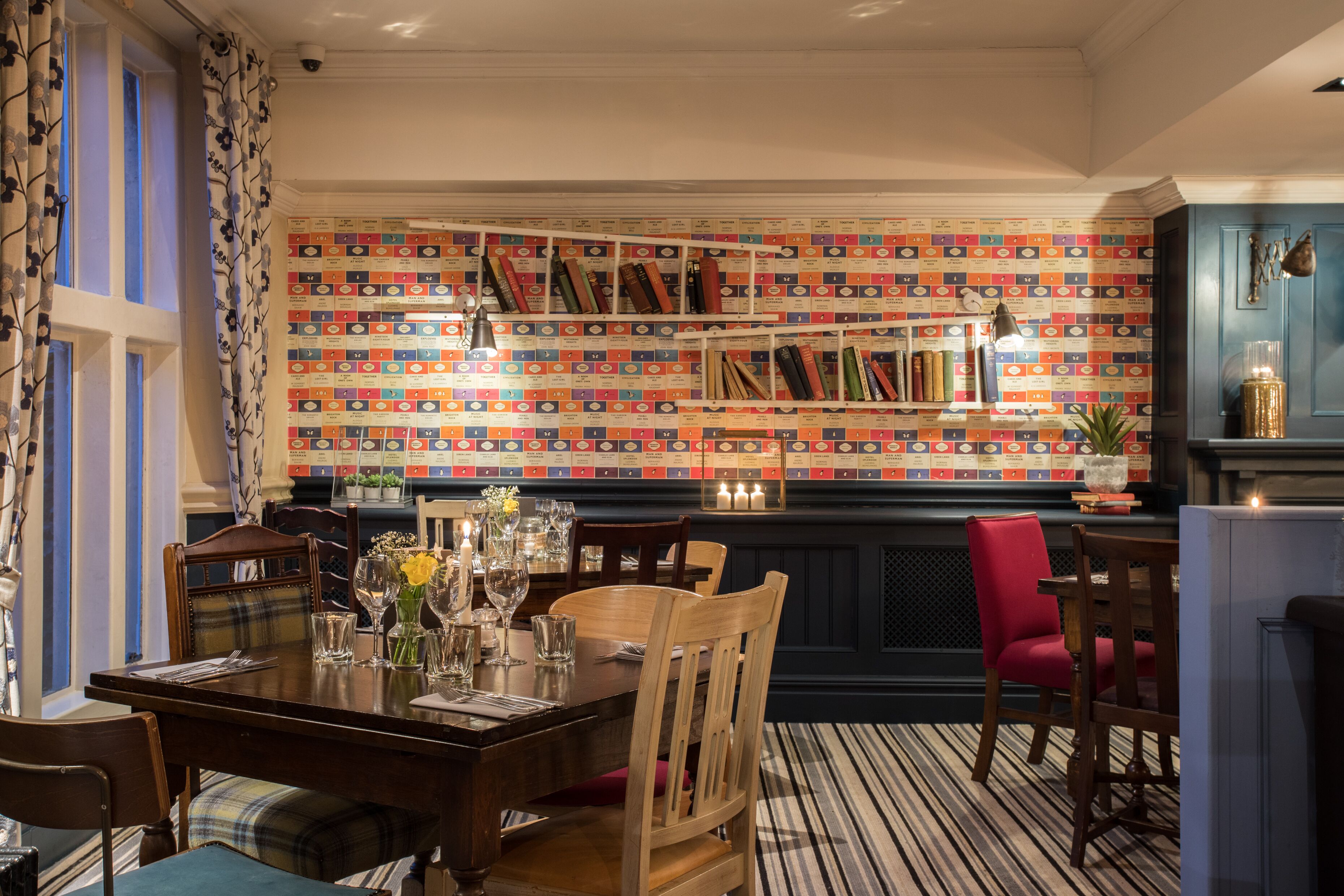 A dining area with a striped floor, wooden chairs, a table set for two, and a bookshelf with books and decorative items.