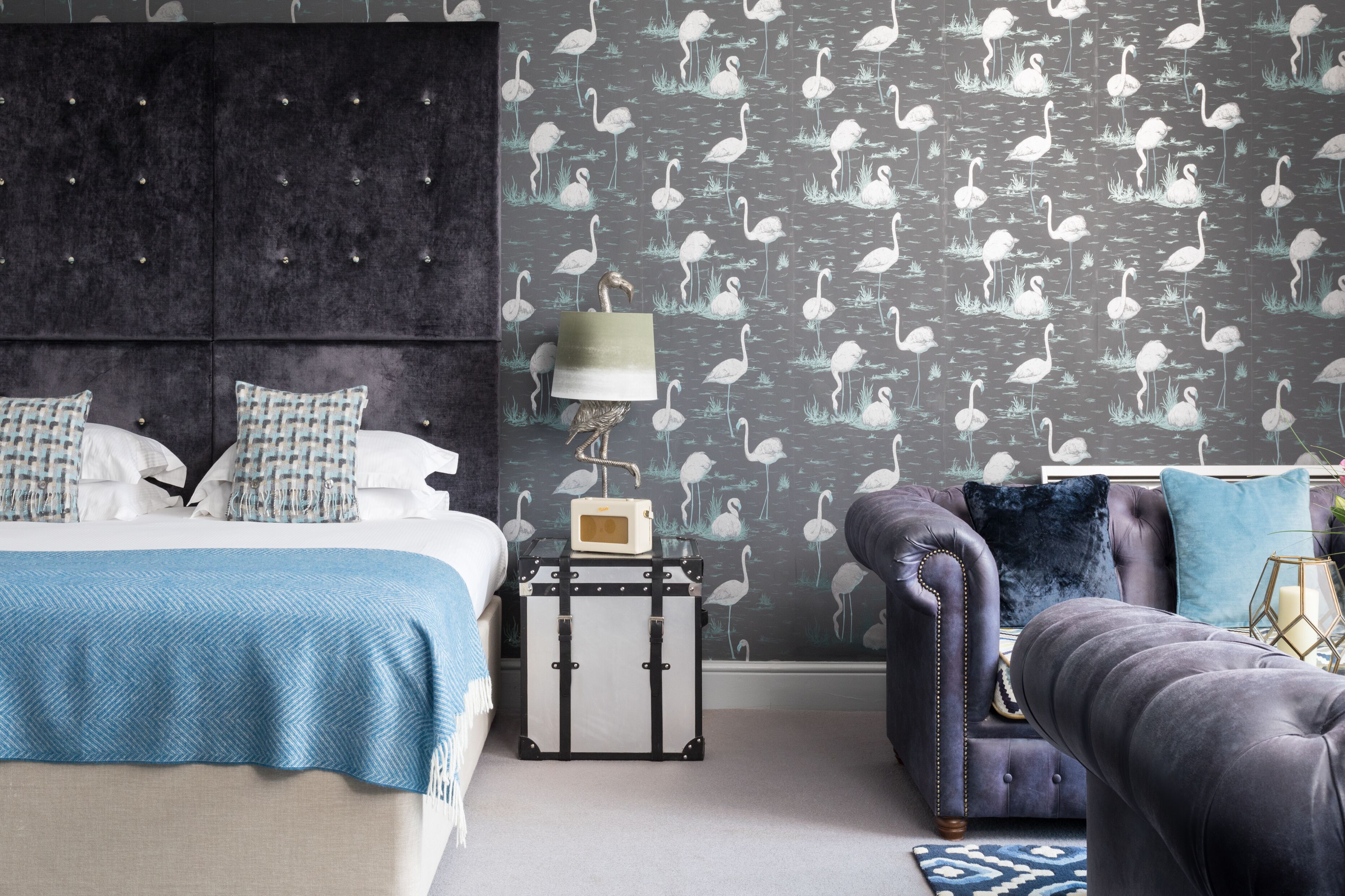 A bedroom with a bed, a dresser, a nightstand, a lamp, and a patterned wallpaper.