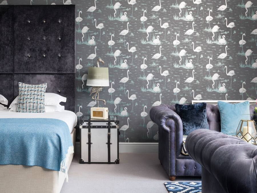 A bedroom with a bed, a dresser, a nightstand, a lamp, and a patterned wallpaper.