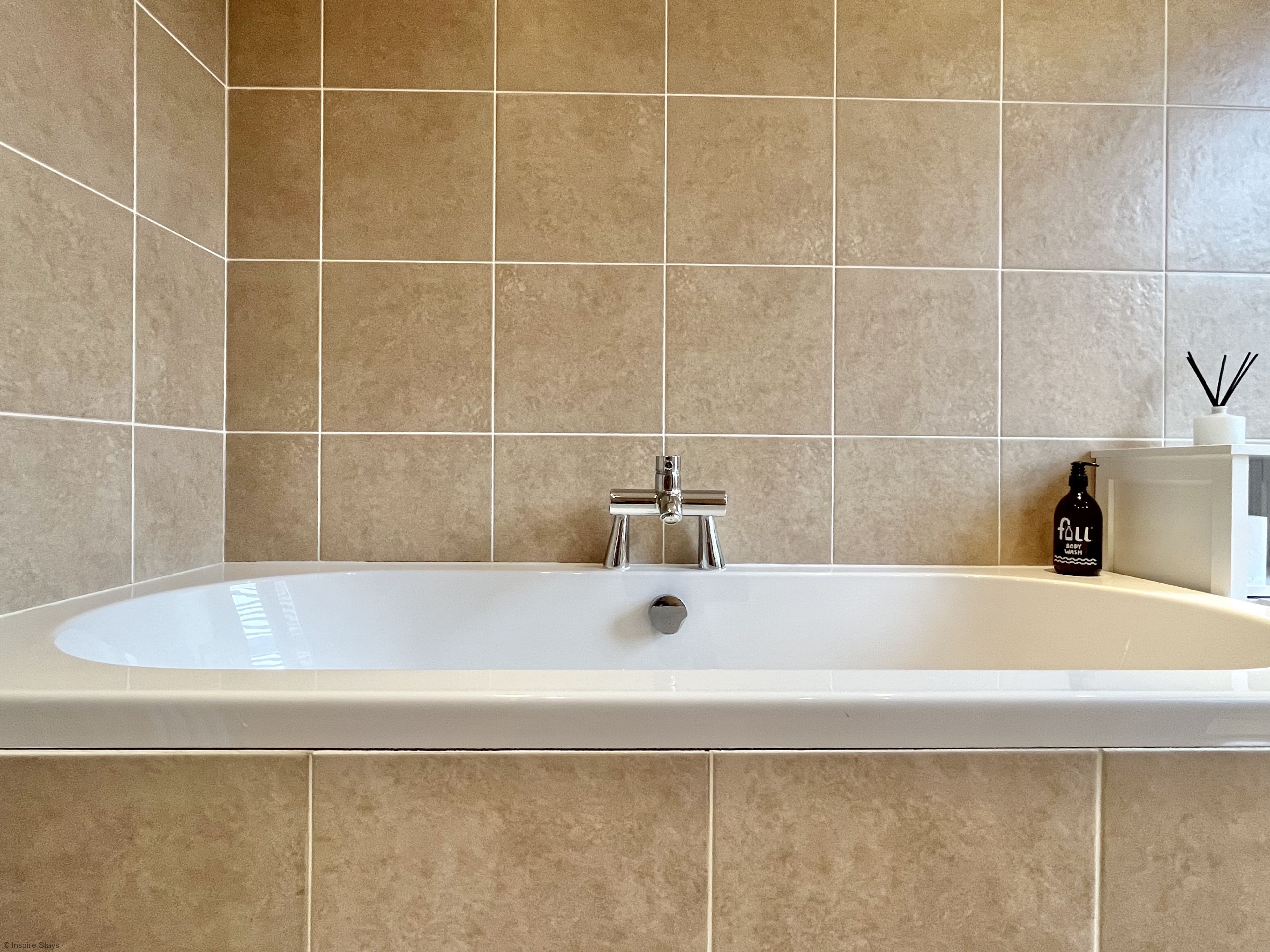 Une salle de bain moderne avec une grande baignoire, des carreaux de couleur beige, un robinet chromé et une bouteille de savon pour la baignoire posée sur le bord.