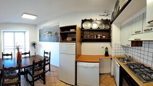 A spacious living area with a blue sofa, a dining table with chairs, a kitchen with a stove, and a TV mounted on the wall. - Azalea Residence (Orta San Giulio)