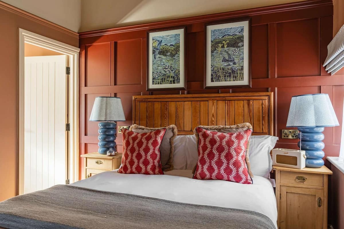A hotel room with a wooden headboard, two framed artworks, a bed with a striped blanket, two bedside tables with lamps, and a wooden nightstand.