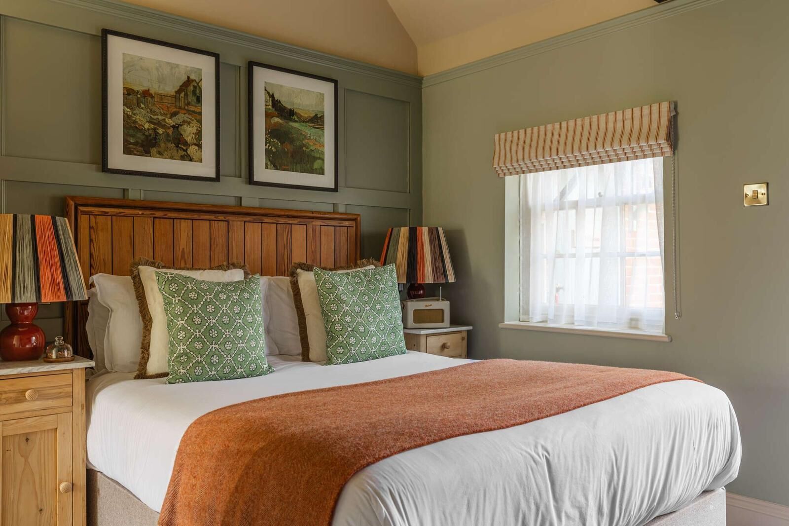 A bedroom with a wooden headboard, a large bed with a white and orange blanket, two green patterned pillows, a nightstand with a lamp, a radio, and a window with a striped valance.