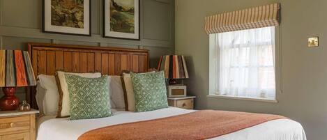 A bedroom with a wooden headboard, a large bed with a white and orange blanket, two green patterned pillows, a nightstand with a lamp, a radio, and a window with a striped valance.