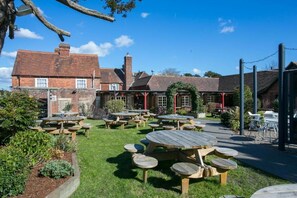 An outdoor seating area with multiple wooden tables and benches, surrounded by a brick building with a garden. - Huntsman of Brockenhurst (Brockenhurst)