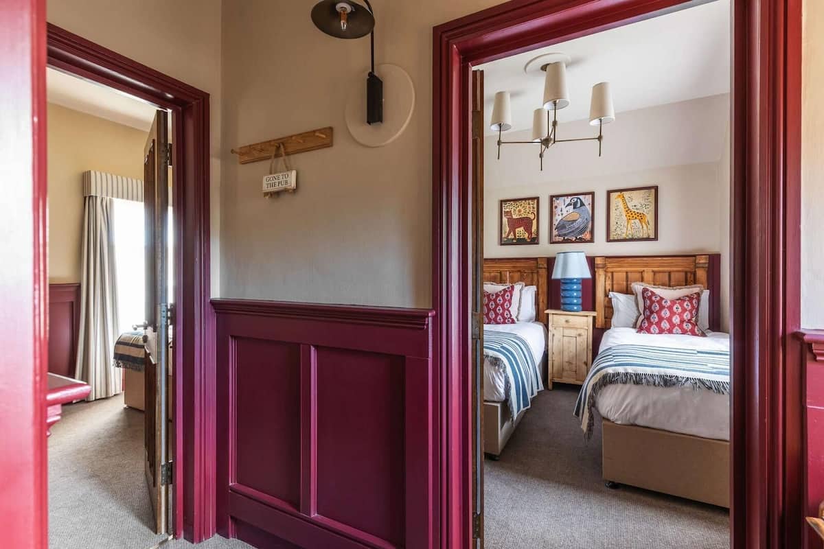 A hotel room with two beds, a wooden headboard, a small table, and a wall-mounted lamp.