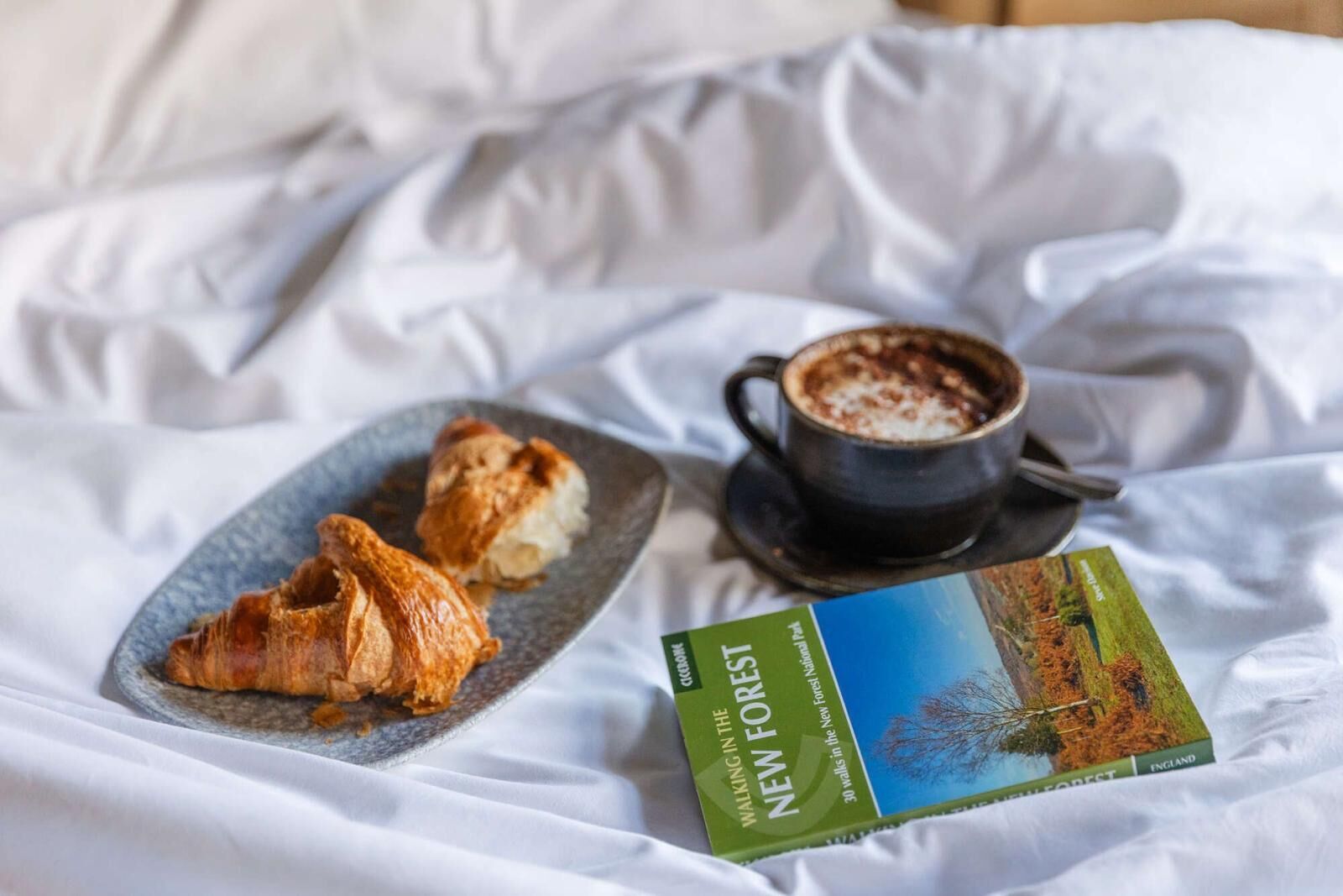 A bed with a plate of croissants, a cup of coffee, and a book titled "Walking in the New Forest".